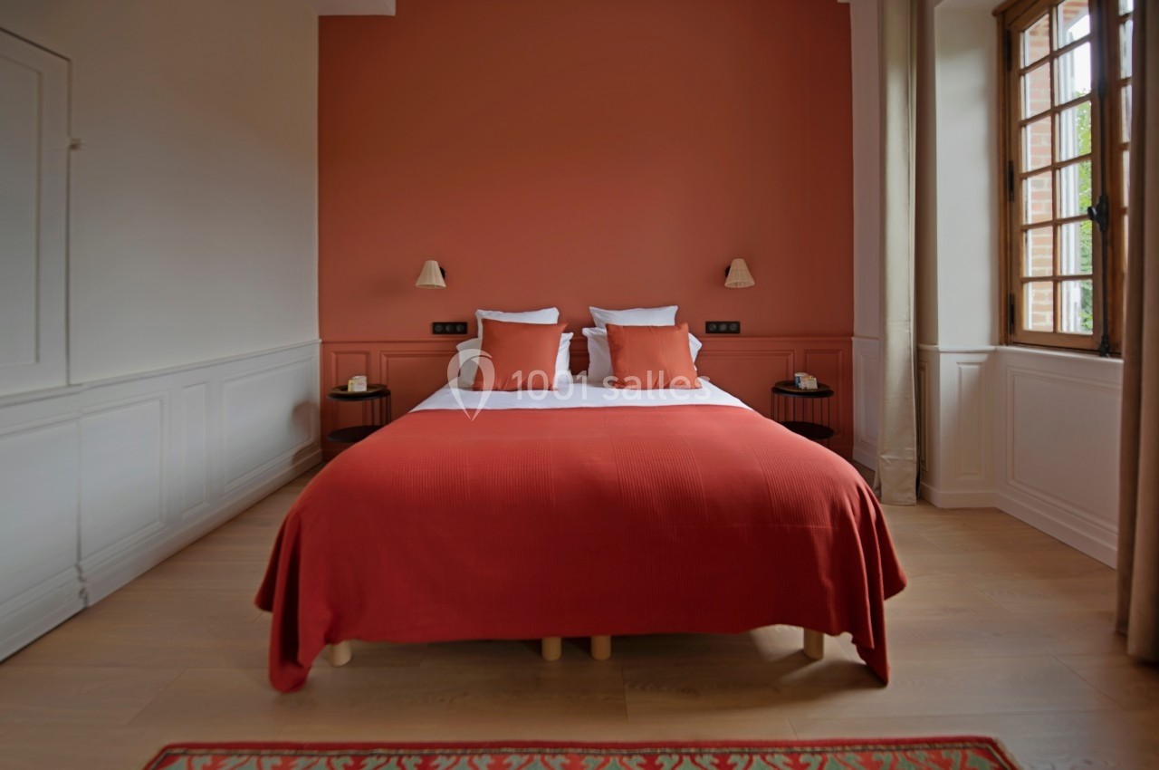 Chambre avec lit double, draps et coussins rouges, murs assortis, éclairage mural et fenêtre avec rideaux beiges.
