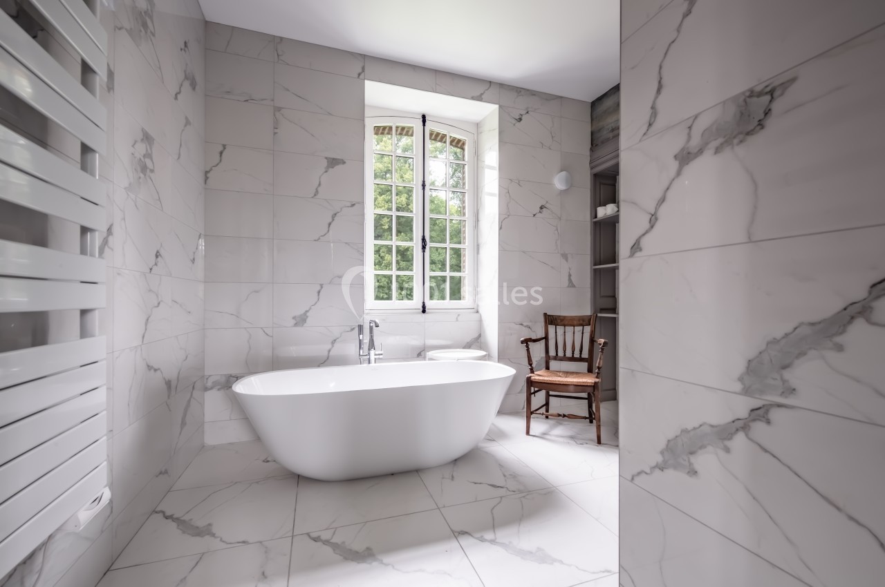 Salle de bain lumineuse avec baignoire îlot, murs et sol en marbre, fenêtre donnant sur un espace vert, chaise en bois.