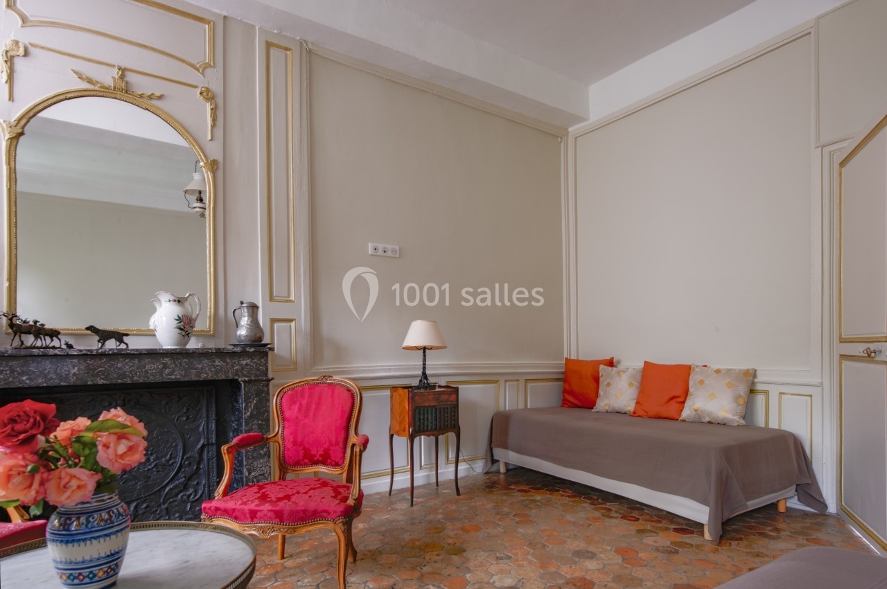 Salon élégant avec moulures, cheminée en marbre noir, fauteuil rouge, canapé beige et table avec vase de fleurs.