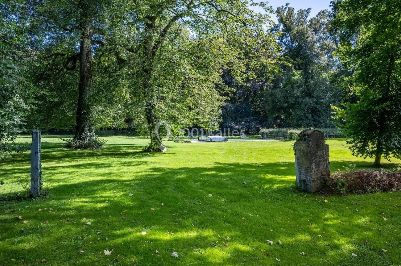 Pelouse verdoyante entourée d'arbres sous un ciel dégagé, avec des pierres anciennes visibles au premier plan.