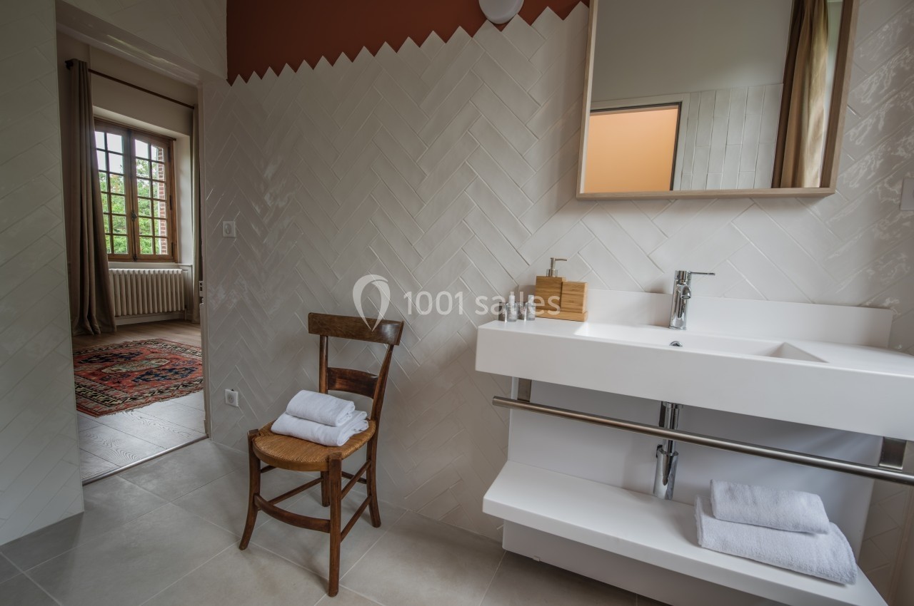 Salle de bain lumineuse avec lavabo moderne, chaise en bois portant des serviettes, et vue sur une pièce adjacente.
