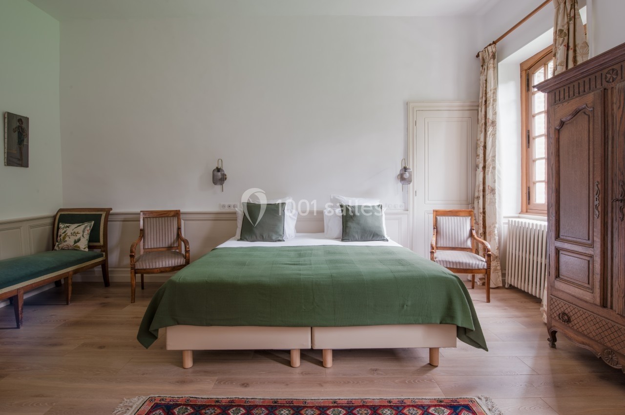Chambre lumineuse avec un lit double, couvre-lit vert, fauteuils en bois, armoire ancienne et tapis coloré.