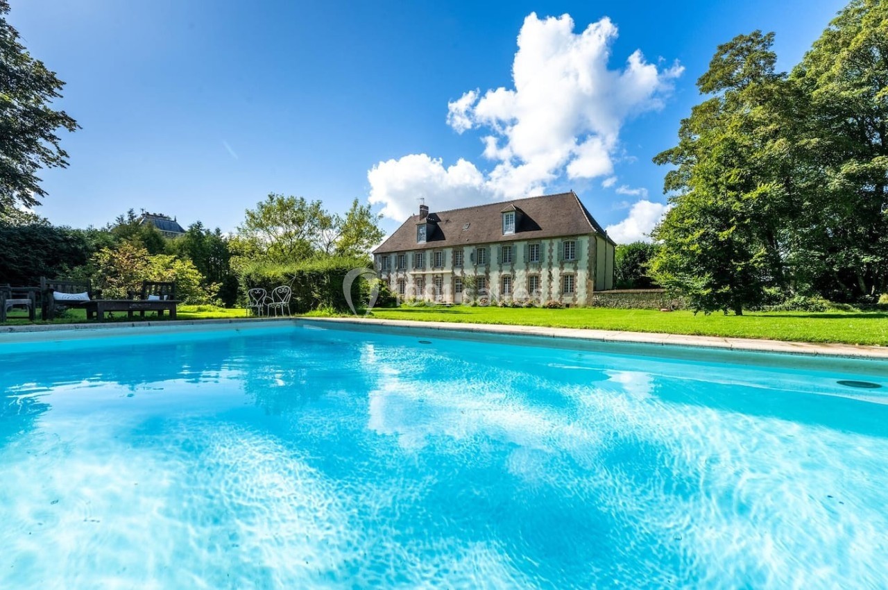 Vue d'une maison de campagne entourée de verdure avec une piscine au premier plan sous un ciel bleu.