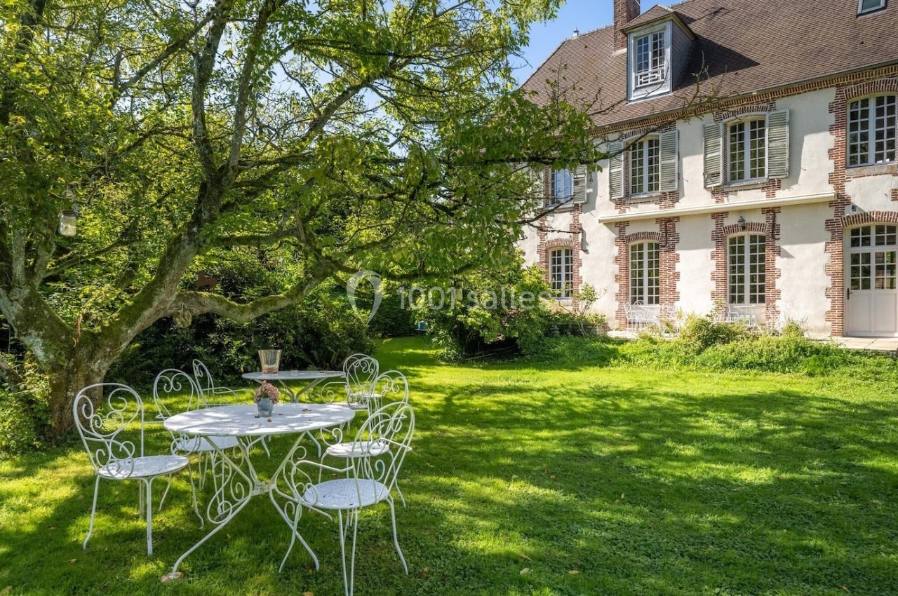 Table et chaises en fer forgé blanc sous un arbre dans un jardin verdoyant, devant une maison en briques et pierres.