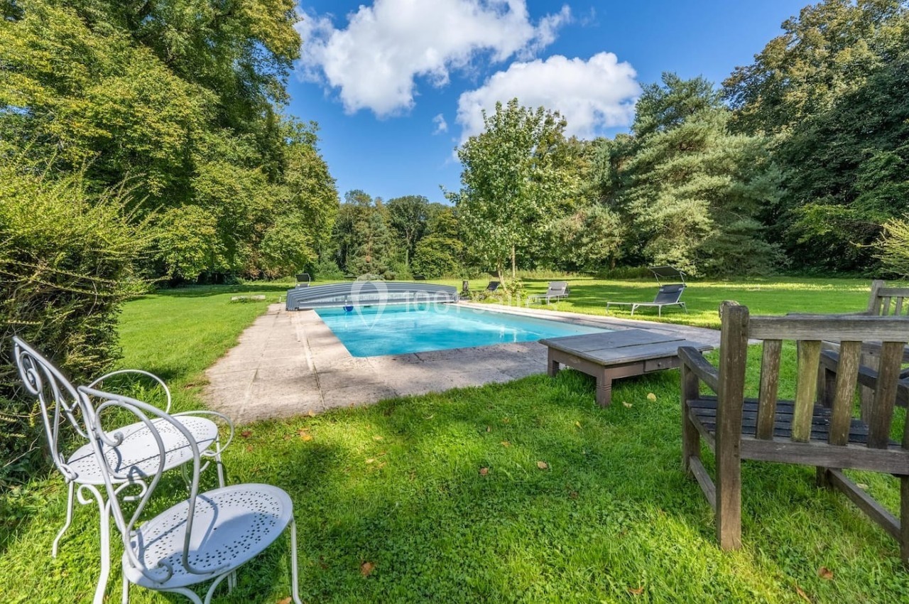 Piscine extérieure entourée de chaises et bancs en métal et bois, située dans un jardin verdoyant sous un ciel bleu.