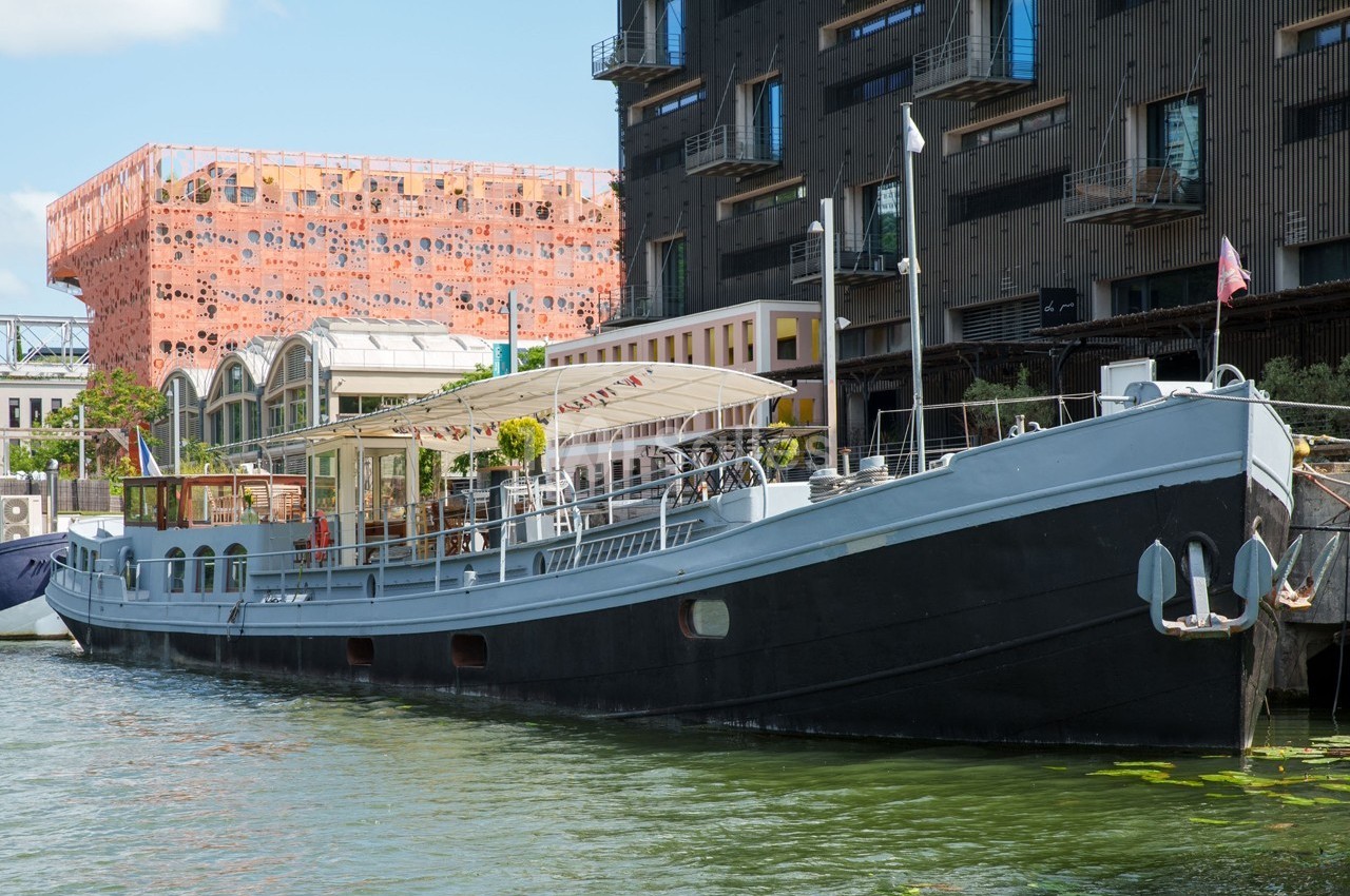 Bateau amarré sur un canal, entouré de bâtiments modernes et d'une structure orange distinctive en arrière-plan.