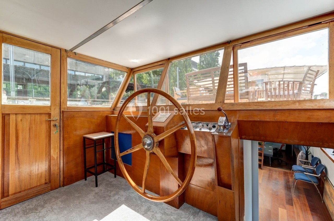 Poste de pilotage en bois d'un bateau avec un grand gouvernail, entouré de fenêtres offrant une vue sur l'extérieur.