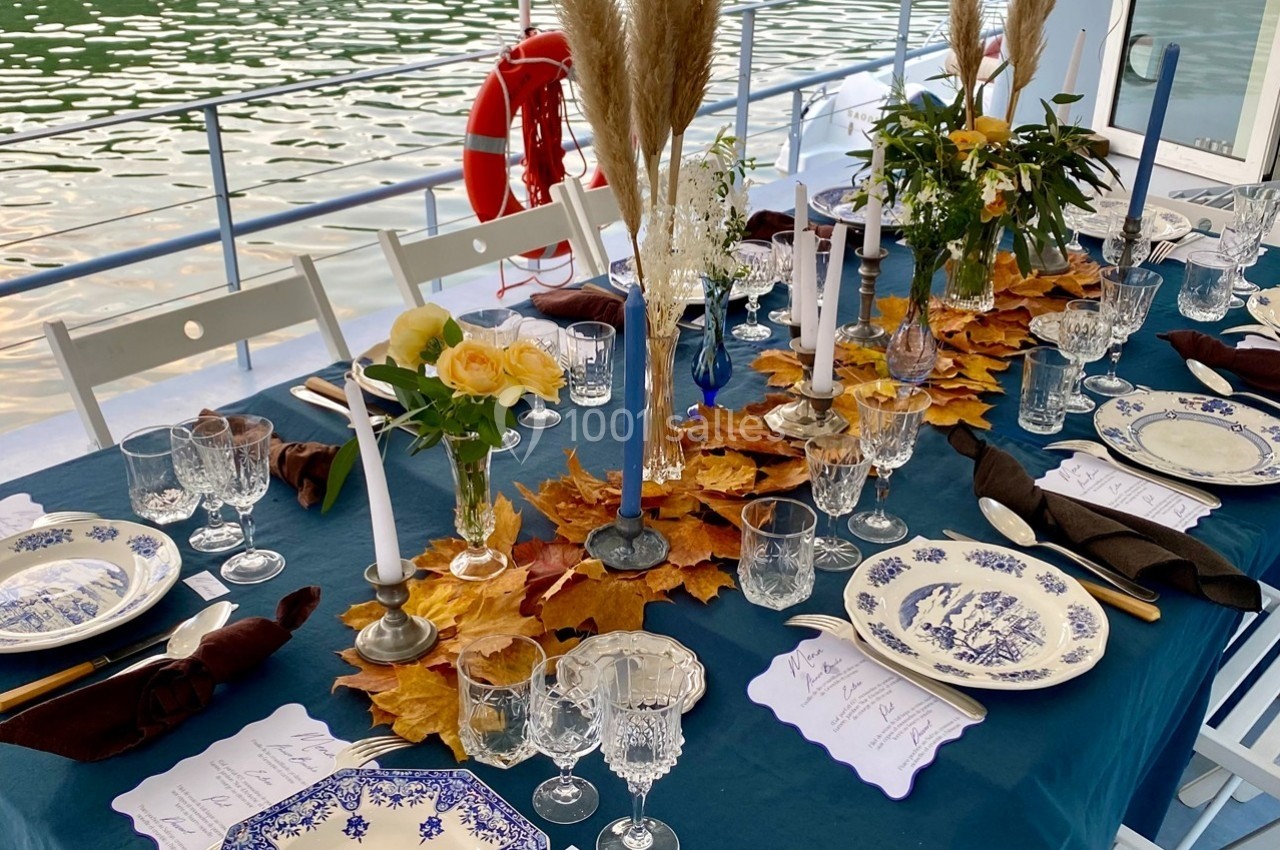 Table élégamment dressée sur une péniche, ornée de feuilles d'automne, bougies et vaisselle en porcelaine bleue.