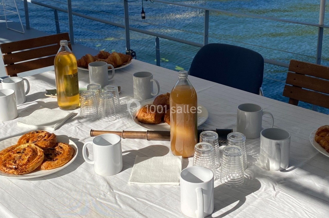 Table dressée en extérieur avec viennoiseries, quiches, bouteilles de jus, tasses et verres, près d'un plan d'eau.