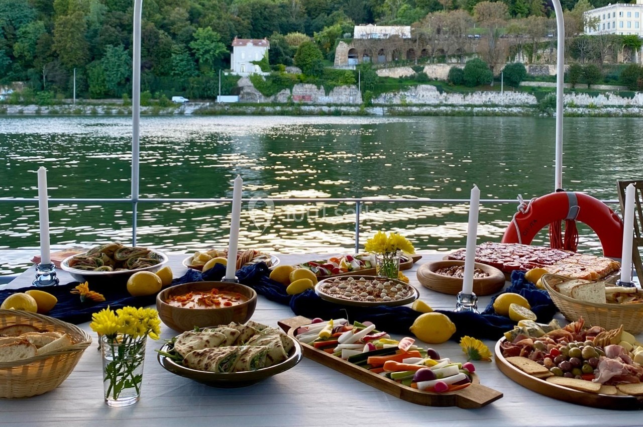 Table dressée avec divers plats et apéritifs, vue sur une rivière et des bâtiments entourés de verdure en arrière-plan.