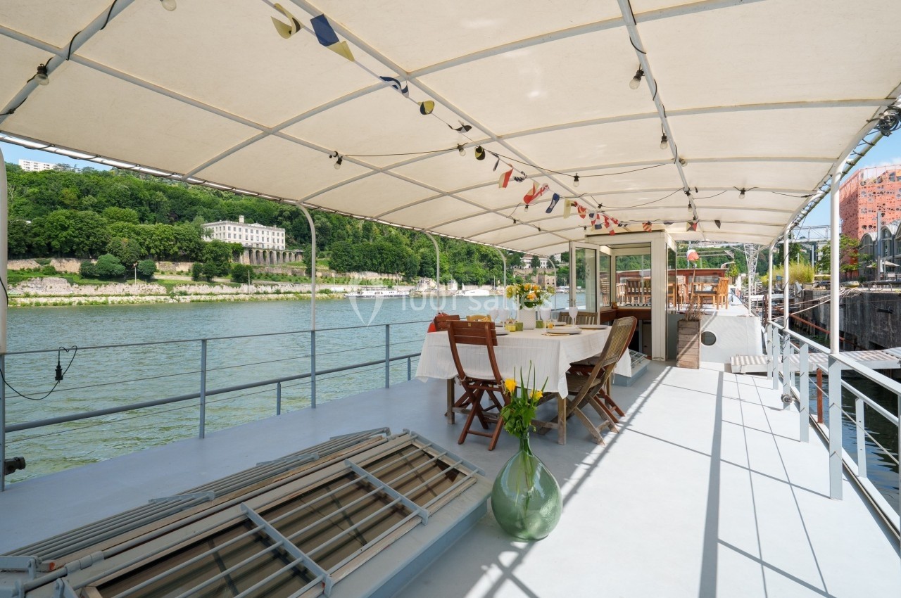 Terrasse couverte d'un bateau avec tables dressées, vue sur un fleuve et des bâtiments entourés de verdure.