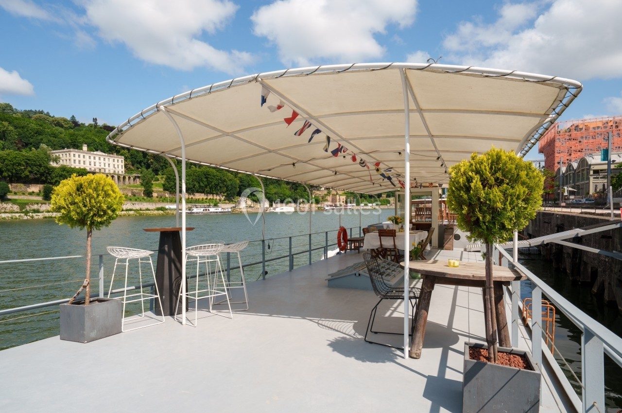 Terrasse d'une péniche aménagée avec des tables, des chaises et des plantes, bordant une rivière sous un ciel dégagé.