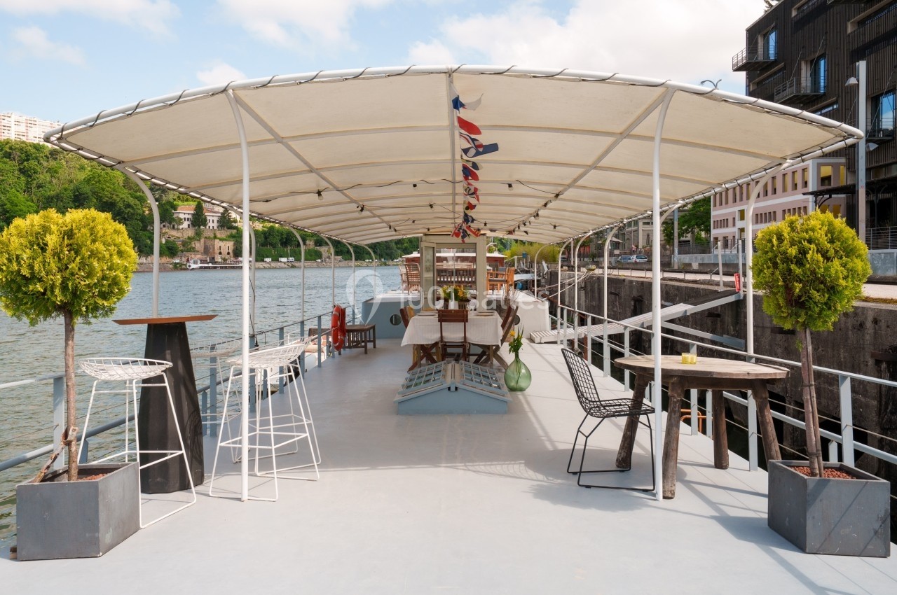 Terrasse d'une péniche aménagée avec des tables, chaises et plantes, amarrée le long d'un quai urbain.