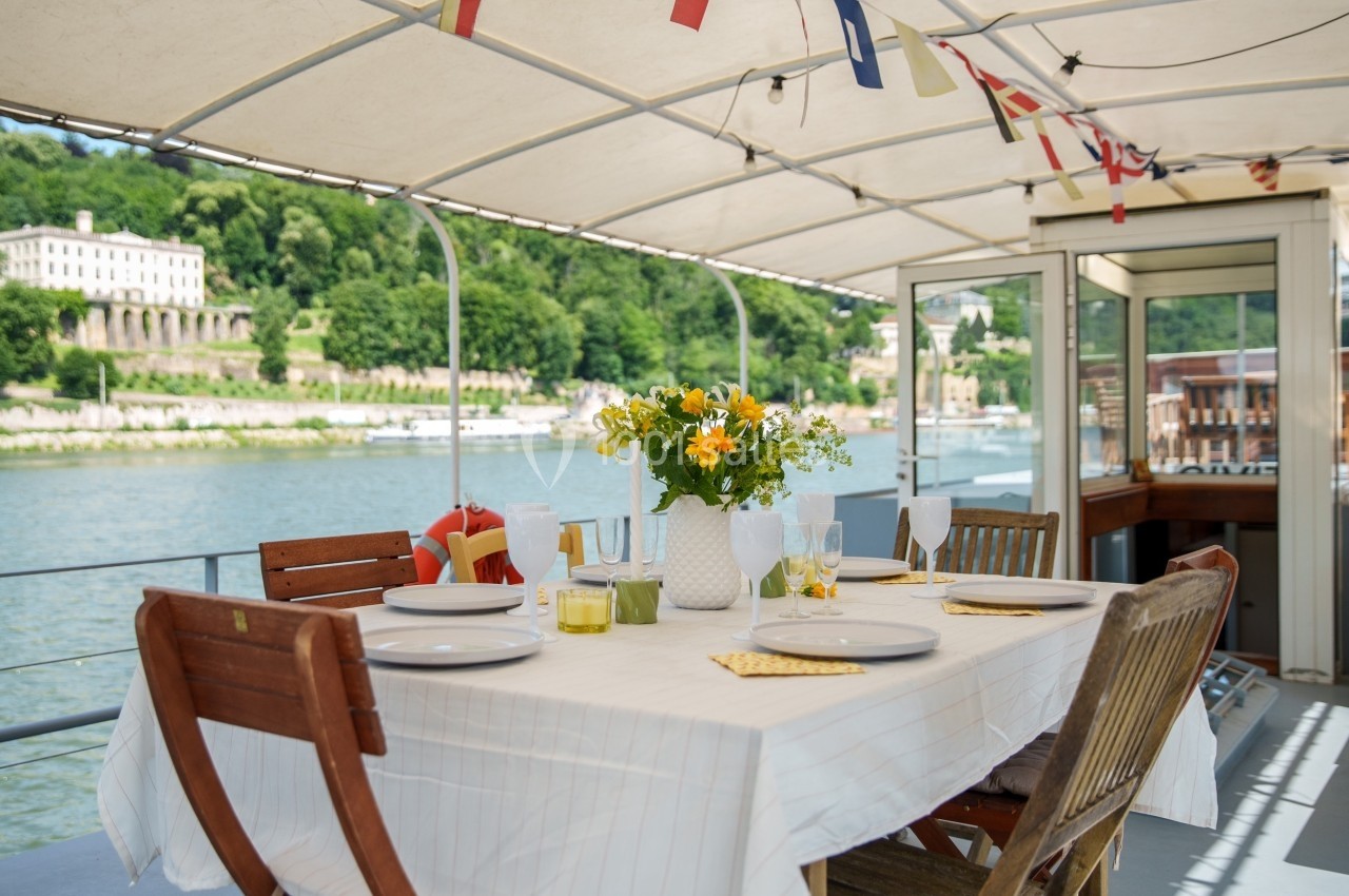 Table dressée avec vaisselle et bouquet de fleurs sur une terrasse couverte, vue sur un fleuve et des collines verdoyantes.