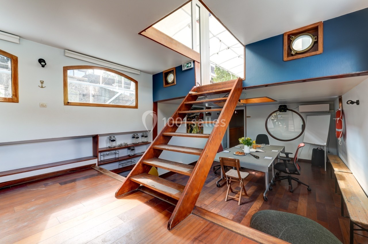 Intérieur lumineux avec un escalier en bois menant à une mezzanine, bureau et table dans un espace aménagé.