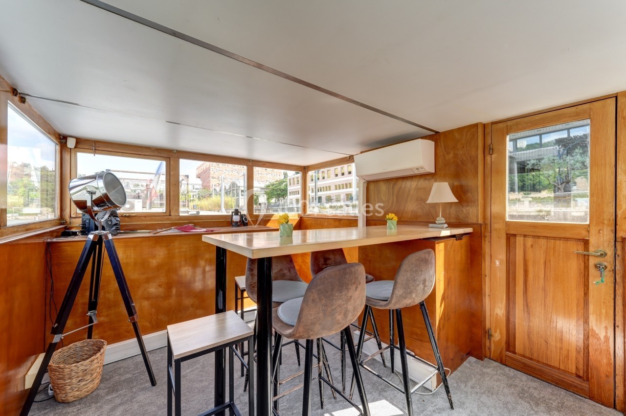 Intérieur lumineux d'une péniche avec table haute, tabourets, télescope et vue sur des bâtiments en arrière-plan.