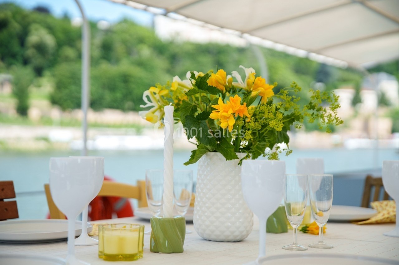 Table dressée en extérieur avec un vase de fleurs jaunes, des verres et des bougies, vue sur un paysage verdoyant.