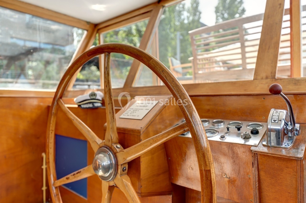 Poste de pilotage en bois d'un bateau avec un grand volant, un tableau de commandes et une vue extérieure floutée.
