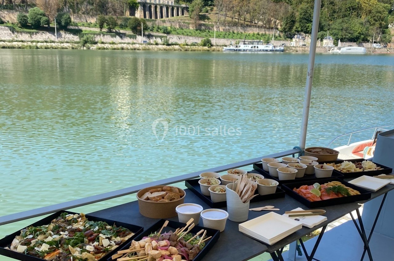 Buffet varié disposé sur une table en extérieur, avec vue sur une rivière et des bâtiments en arrière-plan.
