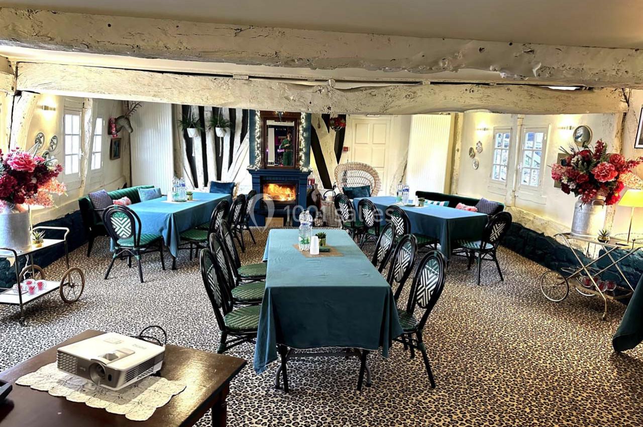 Salle lumineuse avec tables couvertes de nappes bleues, chaises noires, poêle à bois et décorations florales.