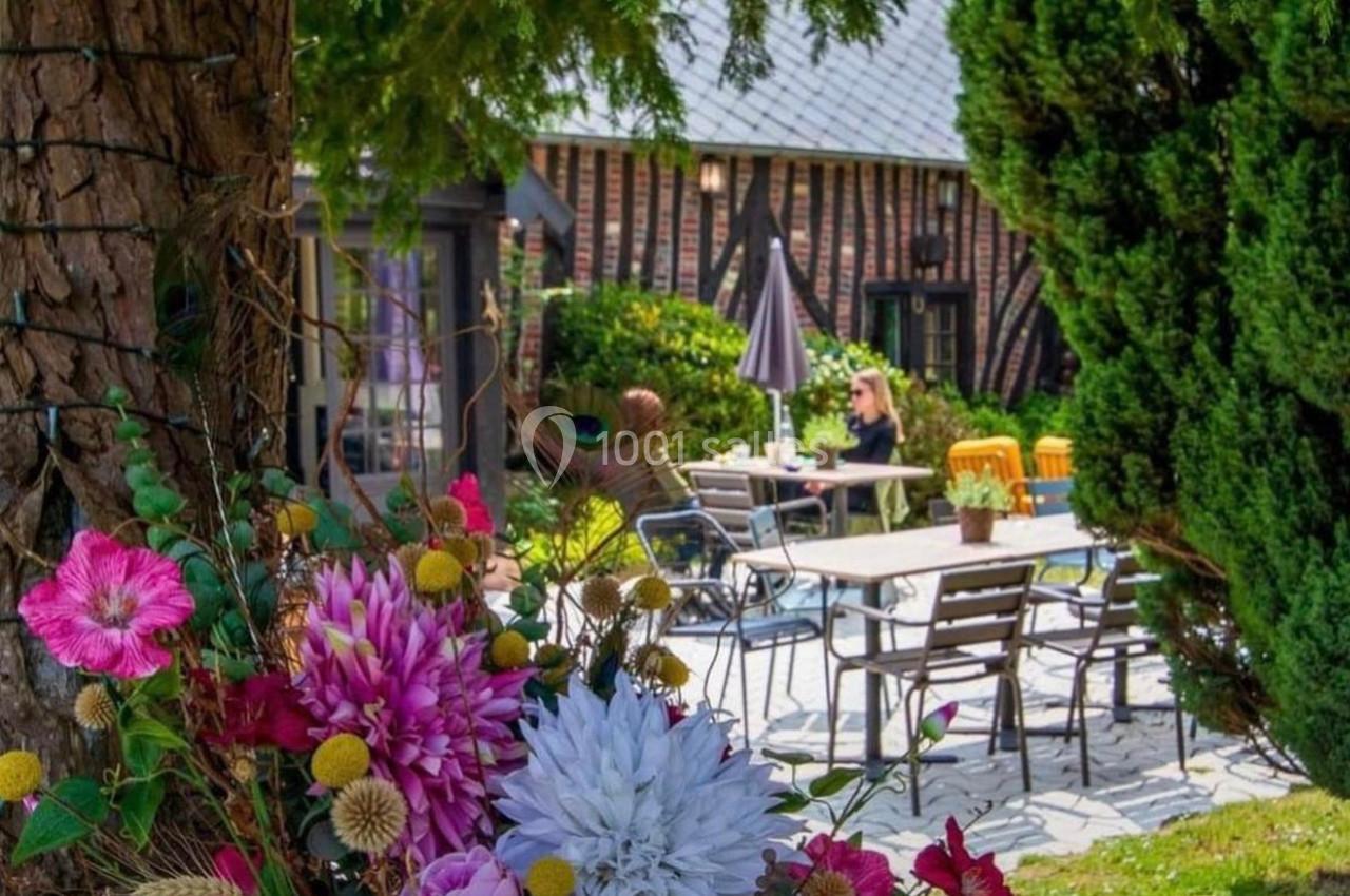 Terrasse extérieure avec tables et chaises, entourée de fleurs colorées et d'arbres, sous un ciel dégagé.