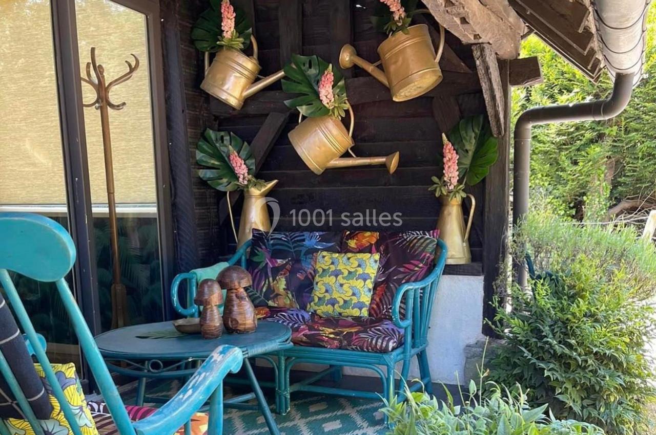 Mobilier de jardin coloré sous une terrasse en bois, décorée d'arrosoirs dorés et de feuillages.