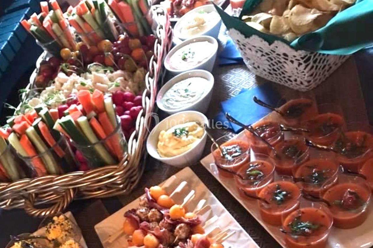 Plateau de buffet avec légumes frais, sauces, chips, brochettes et verrines de soupe disposés sur une table en bois.