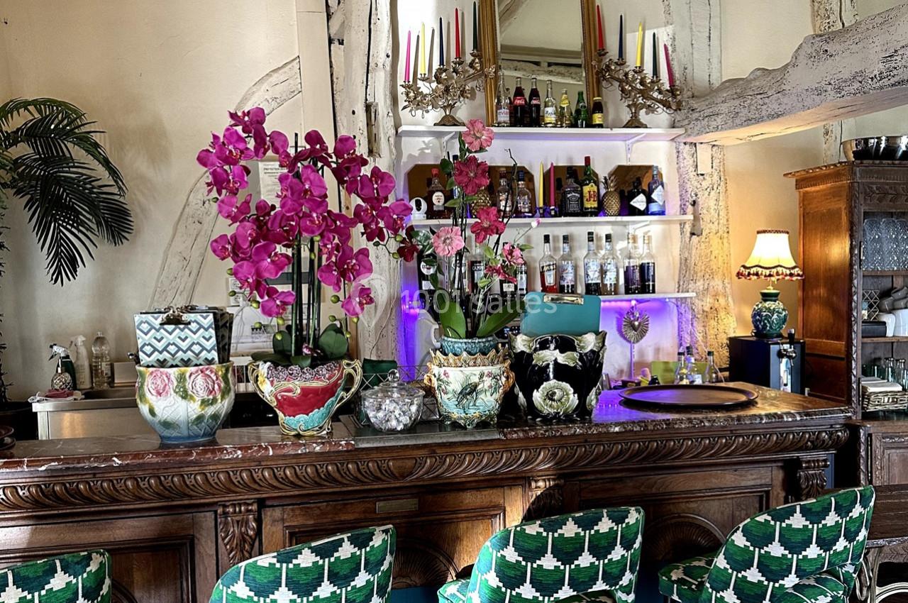 Comptoir en bois décoré de plantes, orchidées roses et pots colorés, avec étagères de bouteilles en arrière-plan.