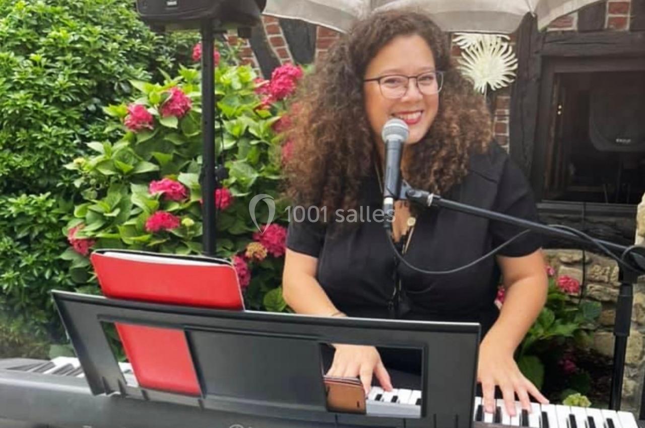 Une femme souriante joue du clavier électronique et chante devant un micro, entourée de fleurs et de verdure.