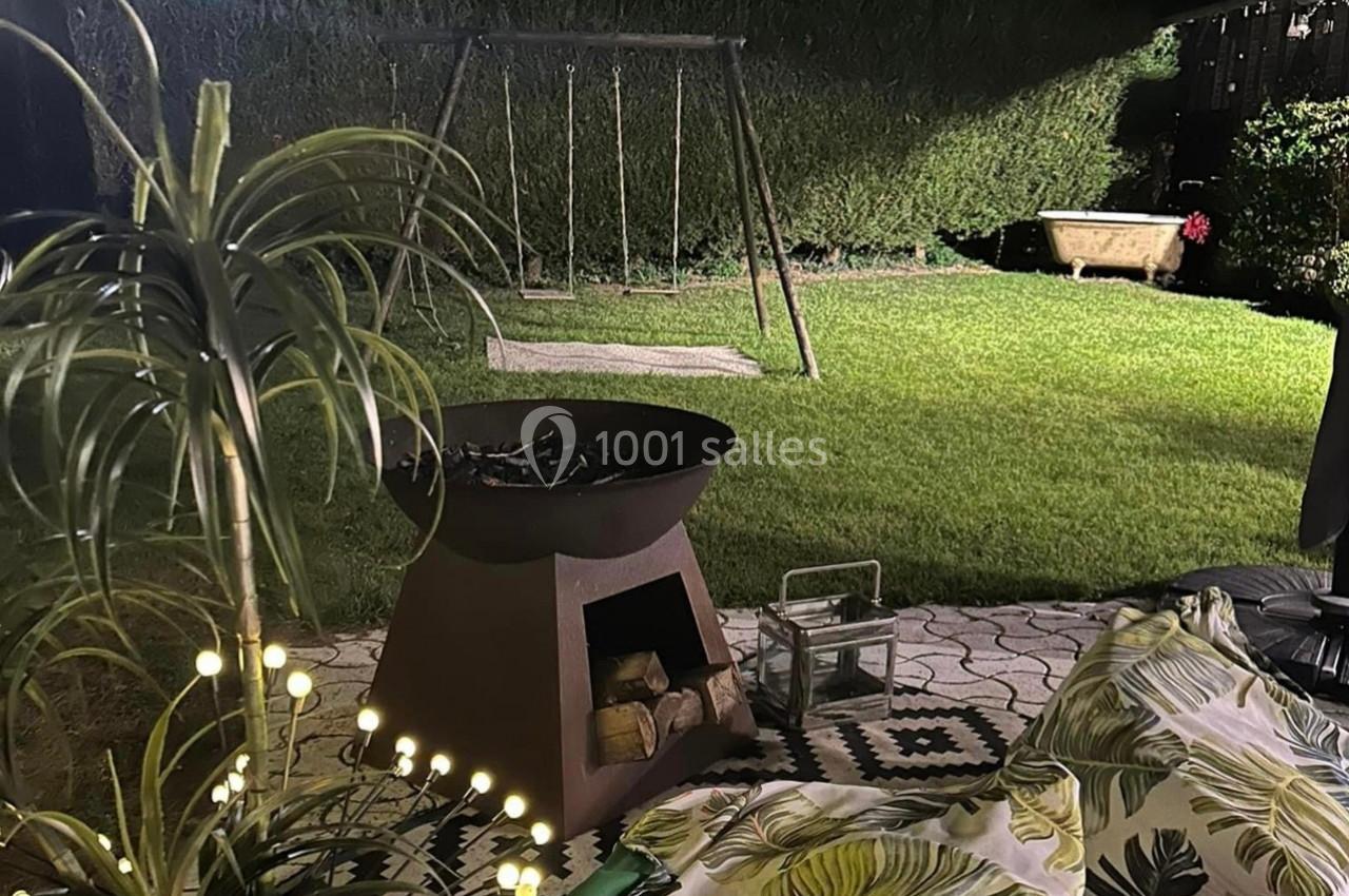 Brasero allumé sur une terrasse éclairée, avec vue sur un jardin, une balançoire et une baignoire ancienne en arrière-plan.