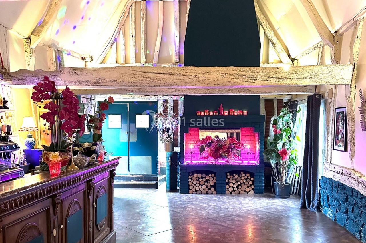 Intérieur d'une salle avec poutres apparentes, cheminée décorée de fleurs et comptoir en bois à gauche.