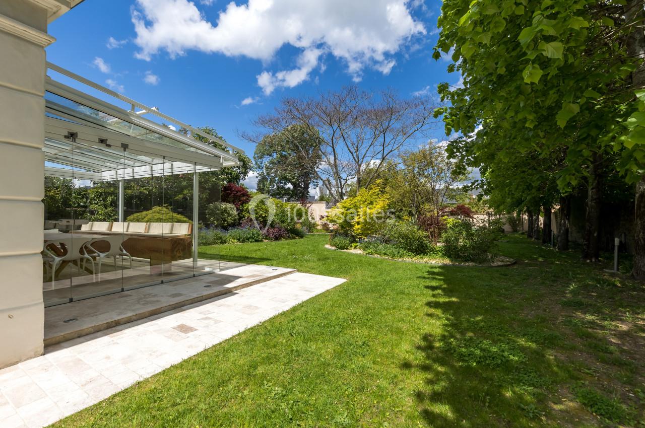 Jardin verdoyant avec pelouse, arbres et arbustes, jouxtant une véranda lumineuse sous un ciel partiellement nuageux.