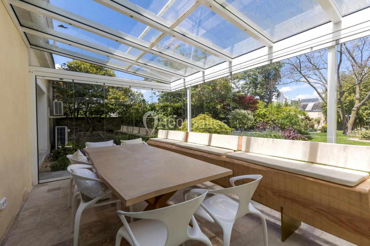 Véranda lumineuse avec toit en verre, table en bois, chaises blanches et vue sur un jardin arboré.