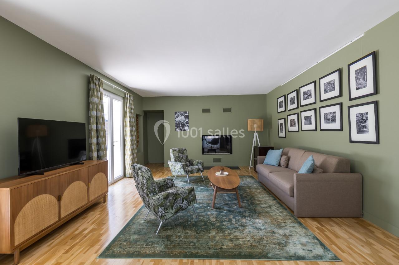 Salon moderne avec murs verts, canapé gris, fauteuils à motifs, tapis bleu, meubles en bois et décorations murales.