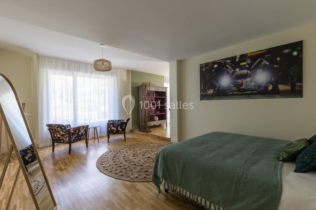 Chambre lumineuse avec lit double, coin salon, miroir sur pied, étagère et grande fenêtre avec rideaux blancs.