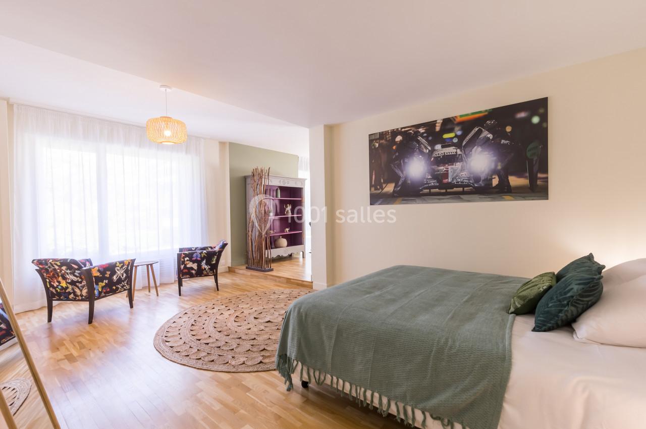 Chambre lumineuse avec un lit double, fauteuils près d'une grande fenêtre, tapis rond et étagère en arrière-plan.