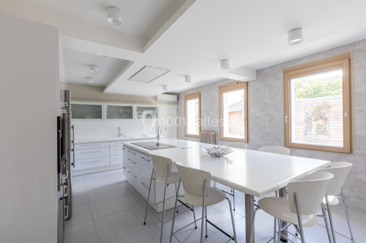 Cuisine moderne lumineuse avec un grand îlot central, des chaises blanches et des fenêtres laissant entrer la lumière…