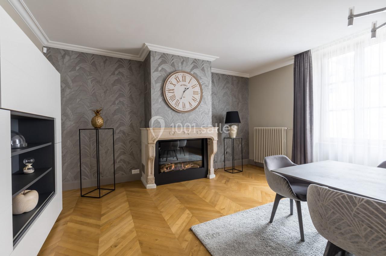 Salon moderne avec parquet en chevrons, cheminée décorative, horloge murale, table à manger et grande fenêtre lumineuse.