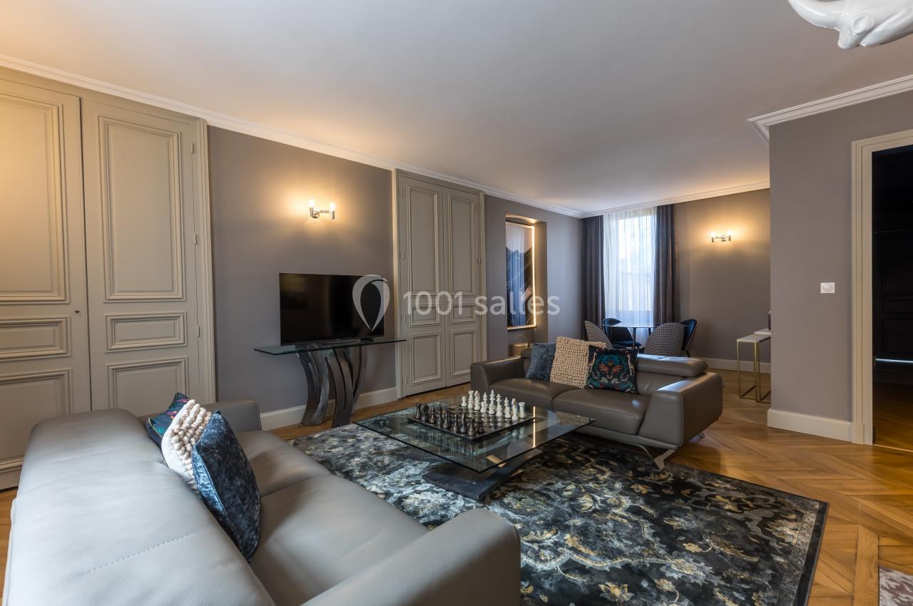 Salon élégant avec canapés gris, table basse avec échiquier, tapis décoratif et éclairage mural doux.