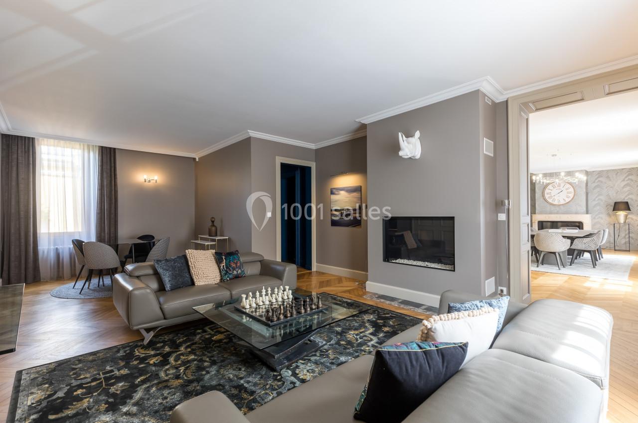 Salon moderne avec canapé gris, table basse en verre, échiquier, cheminée intégrée et vue sur une salle à manger.