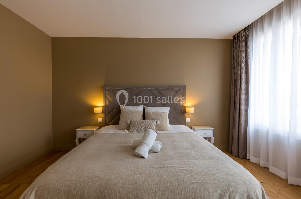 Lit double avec tête de lit capitonnée, draps beiges, deux tables de chevet et lumière naturelle entrant par une fenêtre.