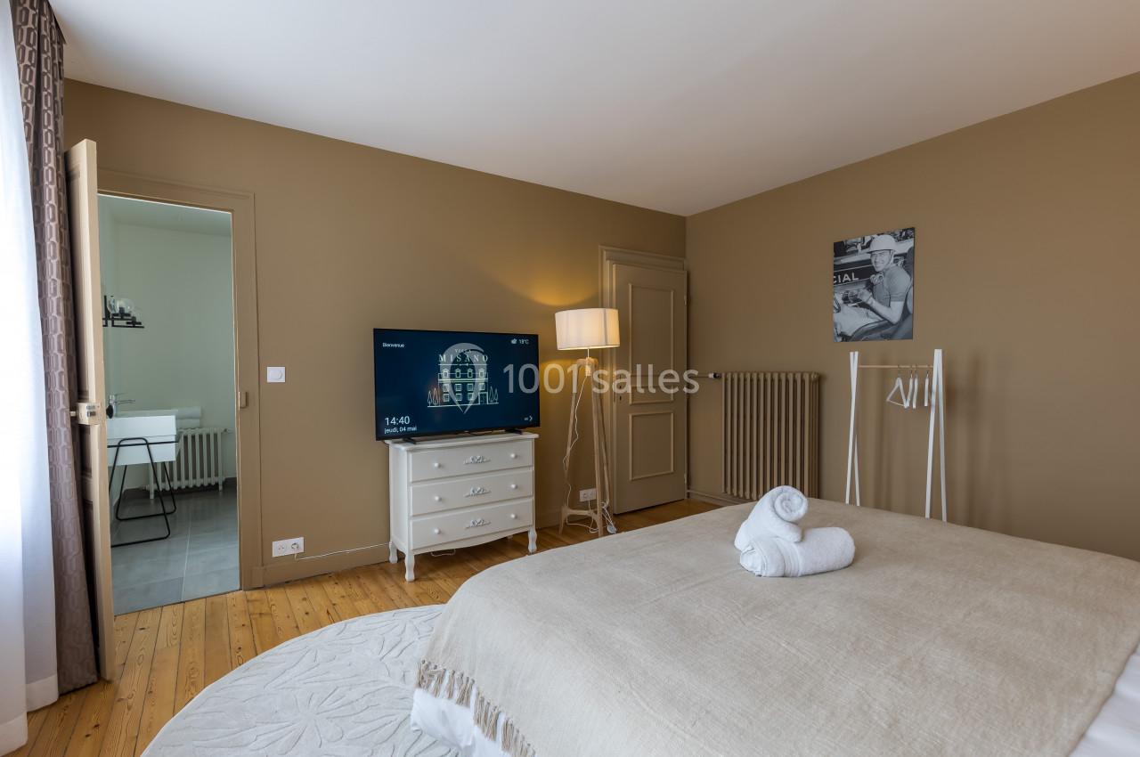 Chambre lumineuse avec lit double, télévision, commode blanche et accès à une salle de bain adjacente.