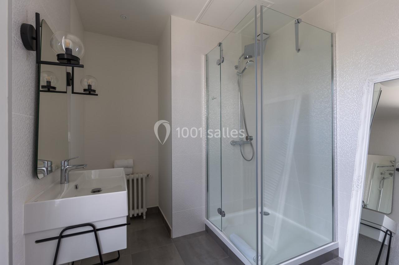 Salle de bain moderne avec douche vitrée, lavabo avec miroir et éclairage mural, murs blancs et sol gris.