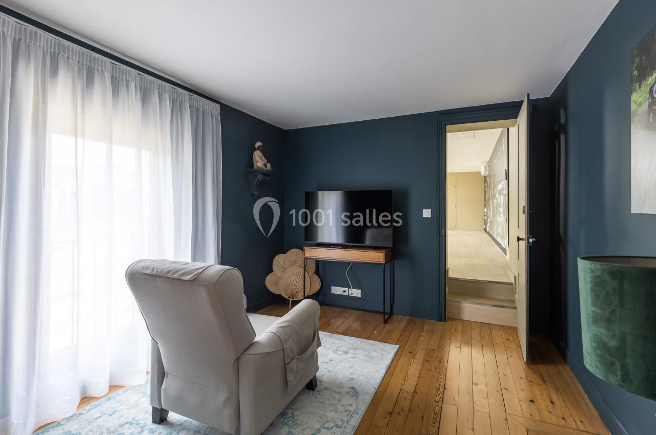 Salon avec fauteuil beige, télévision sur meuble en bois, mur bleu, parquet clair et grande fenêtre avec rideaux blancs.