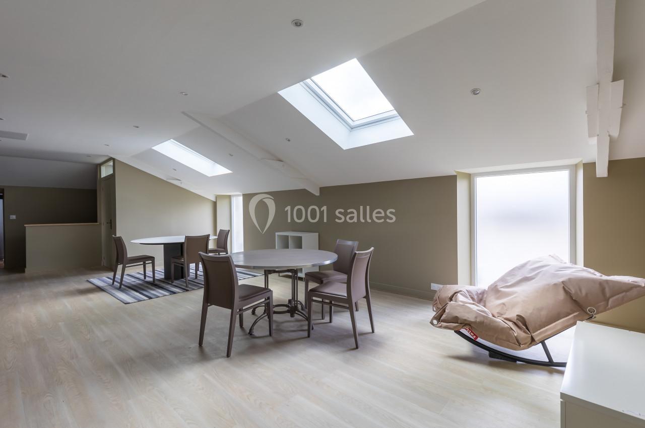 Salle à manger lumineuse sous combles avec table ronde, chaises, fauteuil inclinable et puits de lumière.