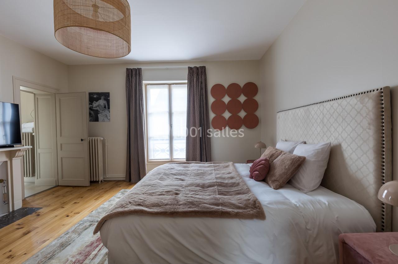 Chambre lumineuse avec un lit double, tête de lit capitonnée, décor minimaliste et fenêtre avec rideaux.