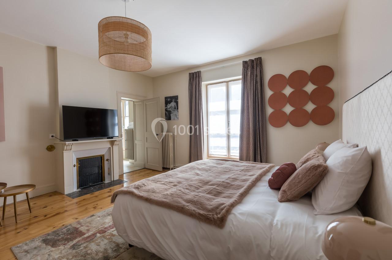 Chambre lumineuse avec lit double, télévision, cheminée, décoration murale et grandes fenêtres avec rideaux.