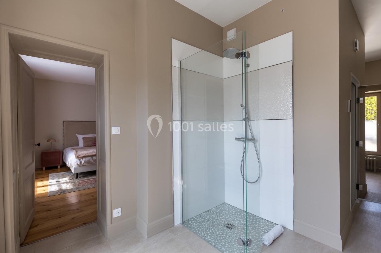 Douche à l'italienne avec parois vitrées dans une salle de bain beige, vue sur une chambre adjacente.