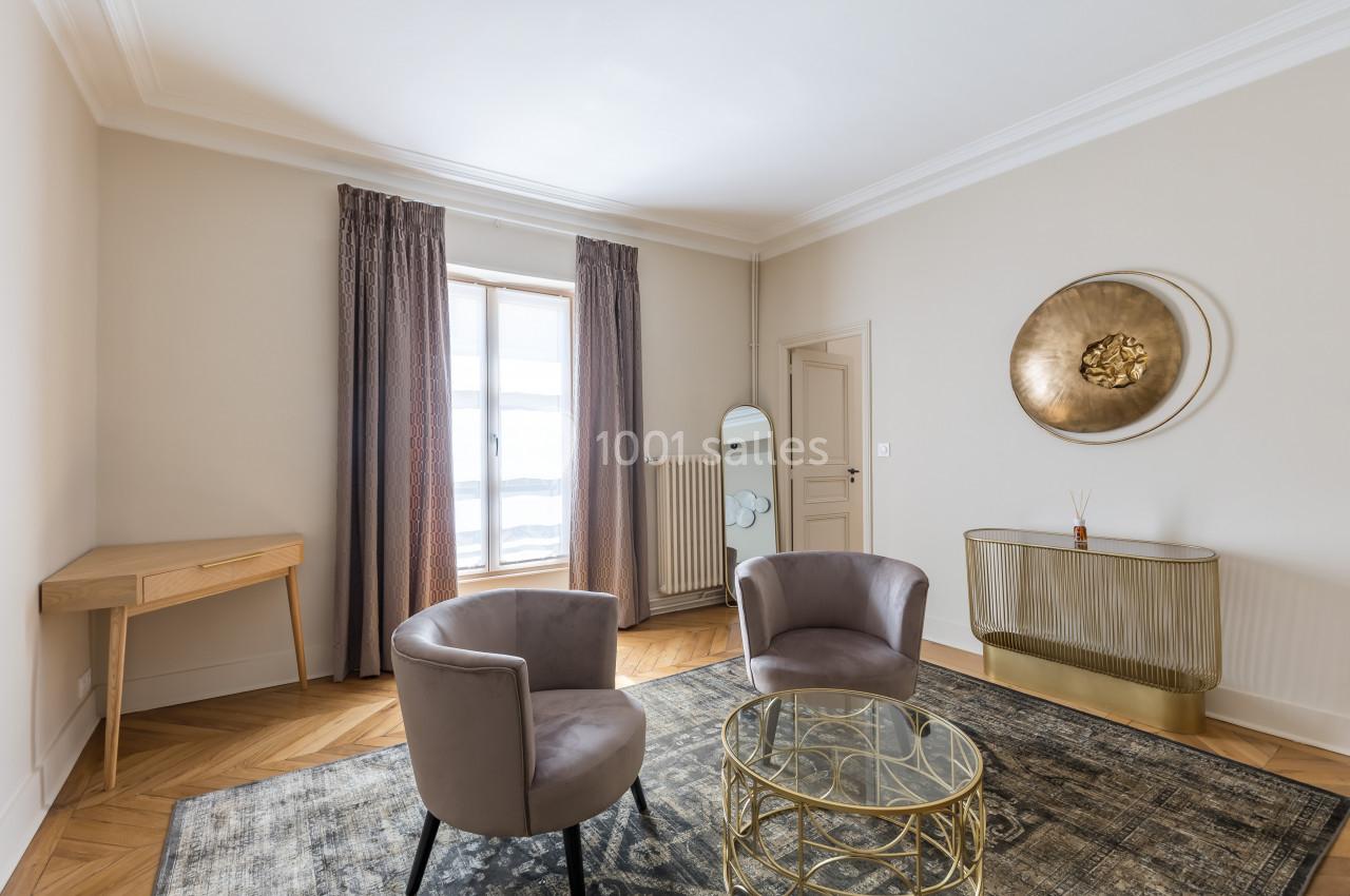 Deux fauteuils gris entourent une table basse en verre dans un salon lumineux avec parquet et décoration murale dorée.