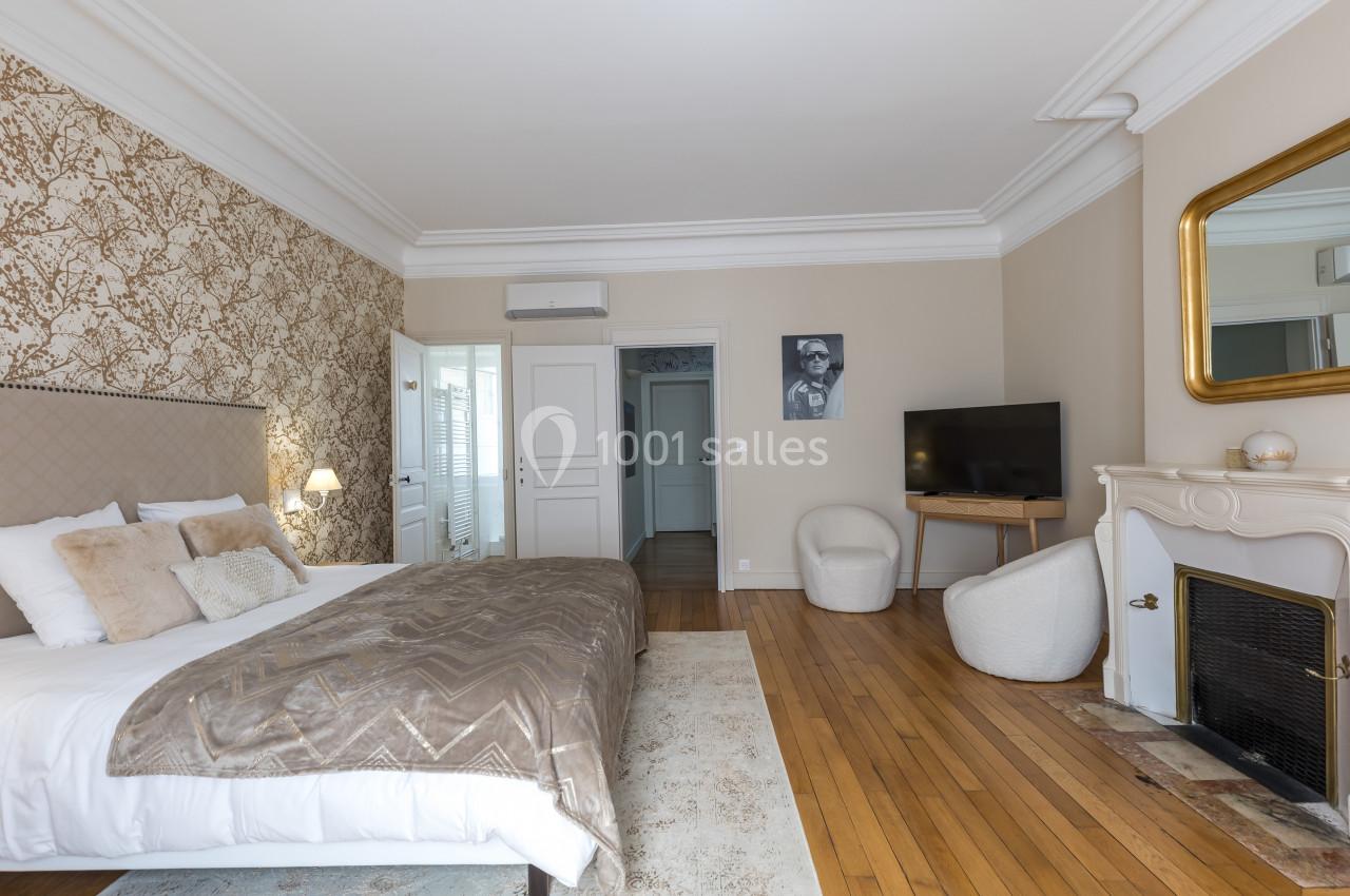 Chambre lumineuse avec lit double, parquet, cheminée, fauteuils, télévision et mur décoré de papier peint floral.